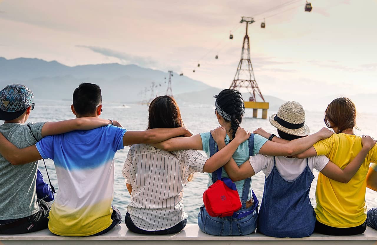 Six people sit in a row with their arms around each other, facing a body of water with cable cars and mountains in the distance, enjoying a scenic view at sunset. Safari Index