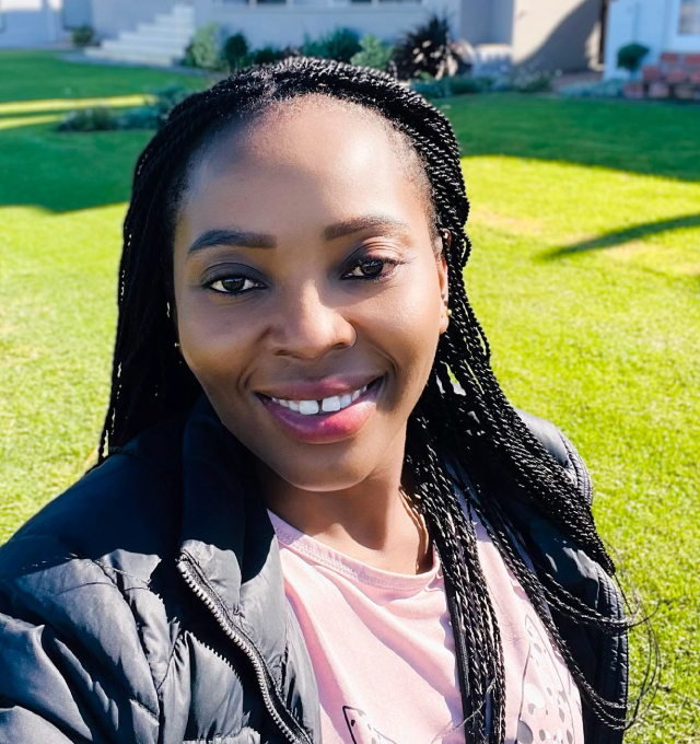 Beauty-Staff-Pic A woman with braided hair smiles at the camera while taking a selfie outdoors on a sunny day, capturing a joyful moment for The Team. She is wearing a pink top and black jacket, with green grass and a building in the background. Safari Index