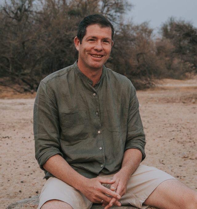 Jake-Hoddinott A man with short dark hair, wearing a green button-up shirt and beige shorts, sits outdoors on a rock, smiling as if hes enjoying time with The Team, with dry trees and sandy ground in the background. Safari Index
